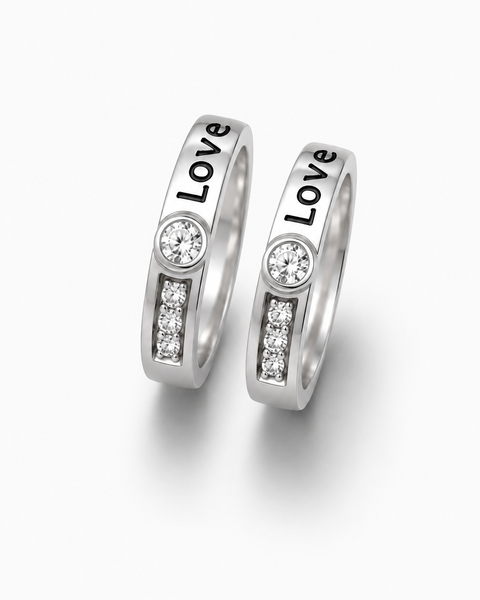 Two silver-toned wedding bands with the word 'Love' engraved vertically, each featuring a round clear gemstone and a small channel of clear stones, shown on a light gray background.