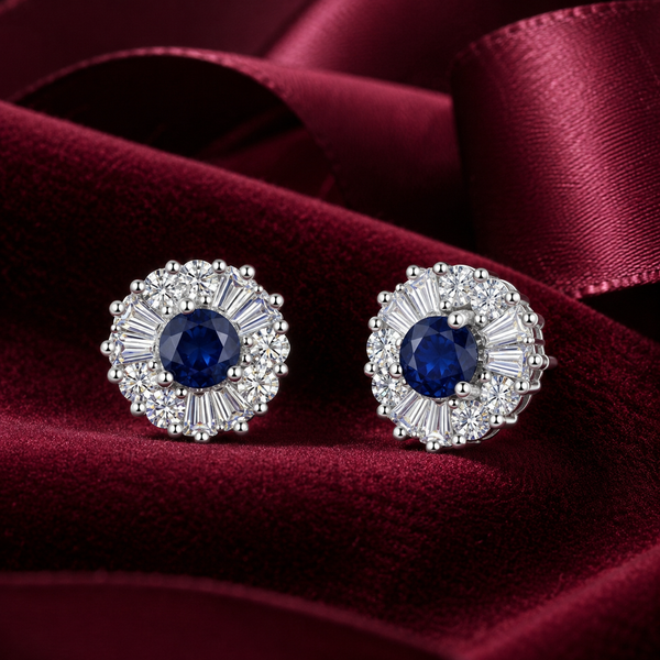 A pair of round stud earrings with a deep blue center stone surrounded by silver-toned baguette and round stones, displayed on a rich burgundy satin fabric.
