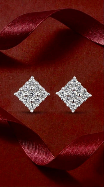A pair of sparkling diamond-shaped stud earrings with a silver-toned halo setting, displayed on a dark red textured background with draped burgundy ribbon accents.
