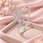 A silver-toned open bangle ring with two flower motifs, one set with clear stones and the other with a pink center stone, displayed in a pink jewelry box on soft pink fabric.