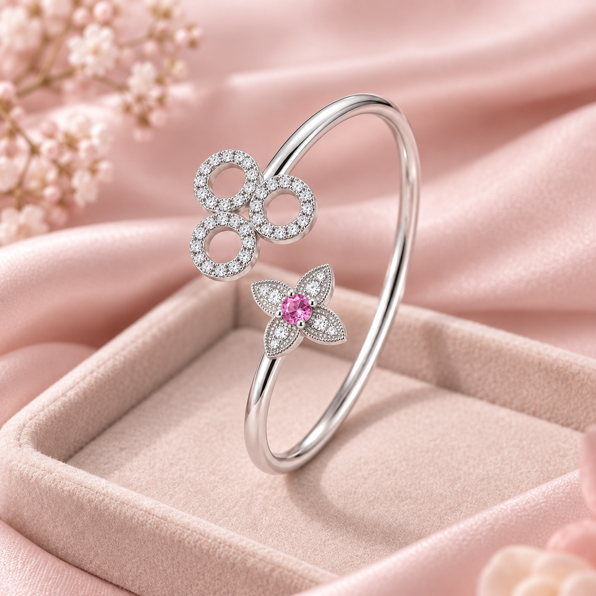 A silver-toned open bangle ring with two flower motifs, one set with clear stones and the other with a pink center stone, displayed in a pink jewelry box on soft pink fabric.