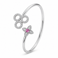 A polished silver-tone open bangle bracelet featuring a large circular floral motif paved with small clear stones and a smaller four-petal flower with a central pink gemstone, shown on a white background.