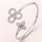A silver-toned hinged bangle bracelet featuring a flower motif with a central pink gemstone and three circular openwork motifs paved with clear stones, set against a soft pink background.