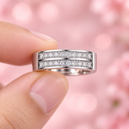 Close-up of a two-row stone-studded silver men’s ring held between fingertips against a soft pink background.