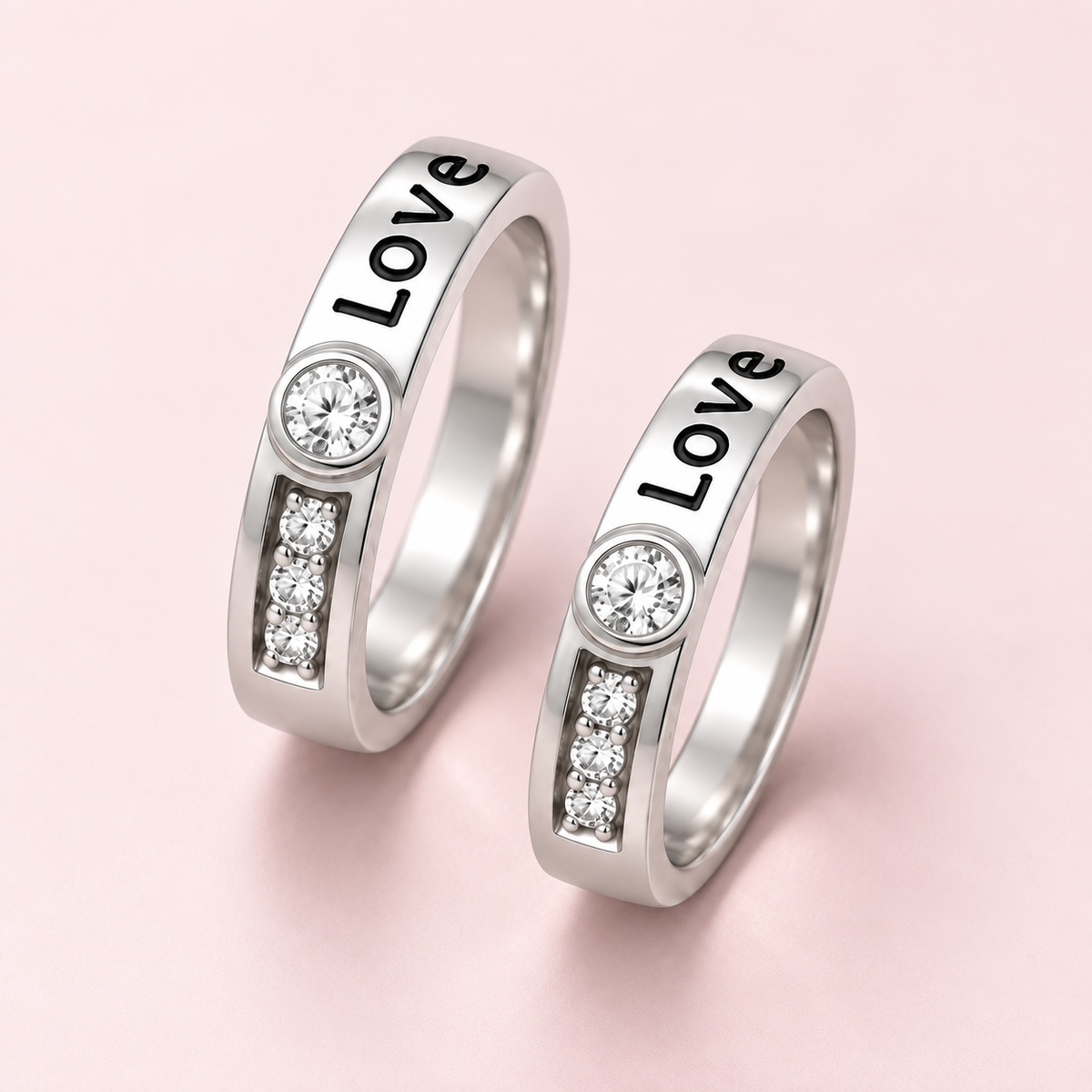 Two silver-toned rings on a soft pink background. Each ring has the word "Love" engraved on the outer band, a round clear gemstone, and a vertical channel of small clear stones.