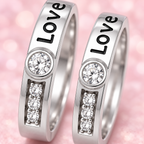 Two silver-toned rings engraved with the word 'Love' in black, each featuring a round clear stone above a vertical row of three clear stones, set against a soft pink bokeh background.