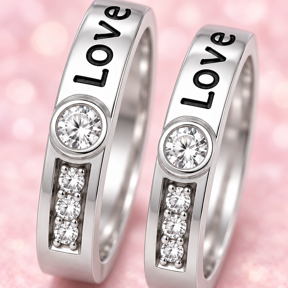 Two silver-toned rings engraved with the word 'Love' in black, each featuring a round clear stone above a vertical row of three clear stones, set against a soft pink bokeh background.