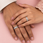 Two hands resting on each other, each wearing a silver band ring engraved with the word 'LOVE' in a delicate script, set against a soft pink background.