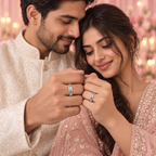 A man and woman in traditional Indian attire holding and looking at silver rings with engraved and stone details, set against a soft pink floral background with warm bokeh lights.