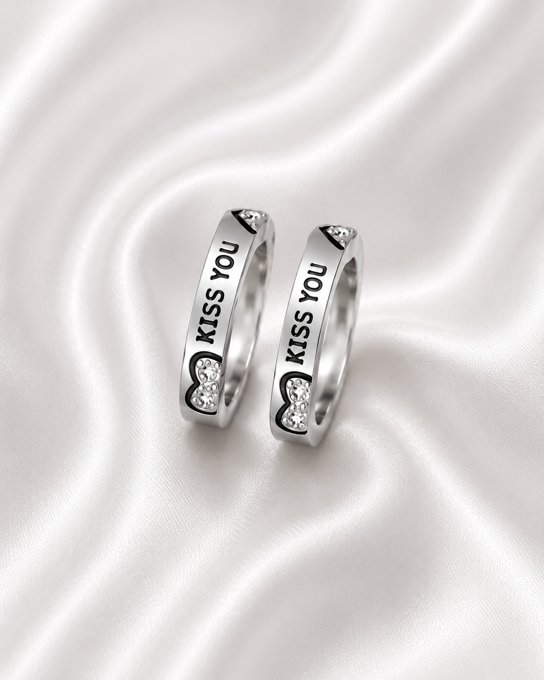 Two silver rings with 'KISS YOU' text and heart-shaped crystal details, displayed on a white satin background.