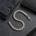 A silver-toned metal link bracelet arranged in an S-shape on a dark gray stone surface, styled with a dark blue shirt cuff and a black leather wallet.