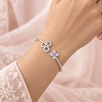 A close-up of a person's wrist wearing a silver bangle bracelet with a three-loop clover design and a smaller flower motif with a pink center, set against a soft pink fabric background.