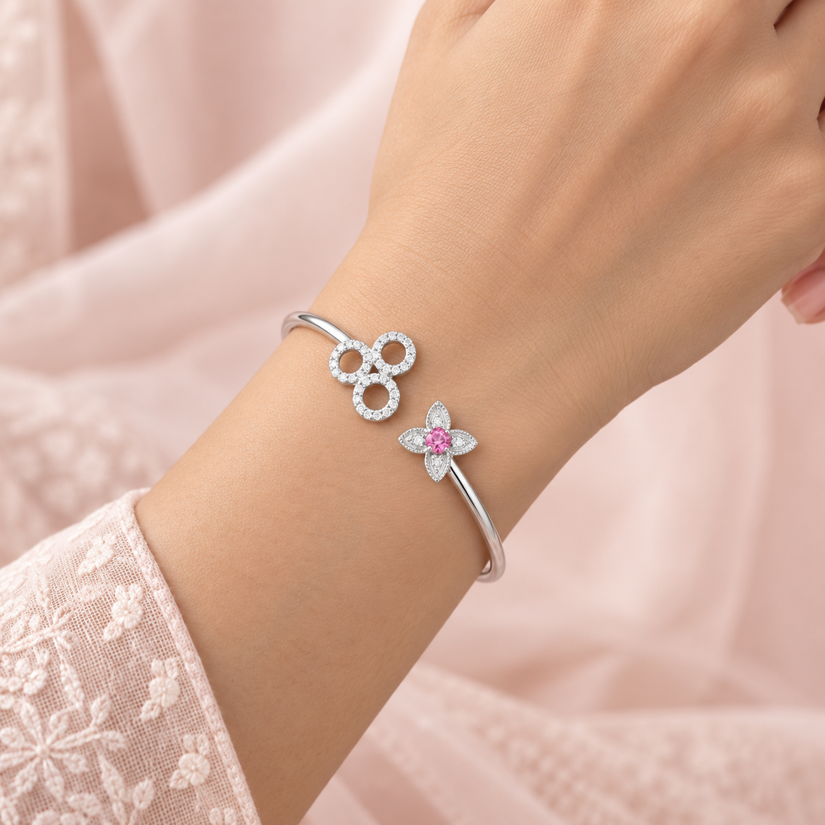 A close-up of a person's wrist wearing a silver bangle bracelet with a three-loop clover design and a smaller flower motif with a pink center, set against a soft pink fabric background.