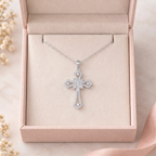 A silver cross pendant necklace with clear stones, displayed inside an open light pink jewelry box on a soft pink ribbon, with small dried flowers in the corner.