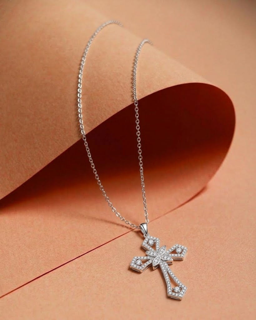 A silver cross pendant necklace paved with small clear stones, resting on a peach-colored paper background.